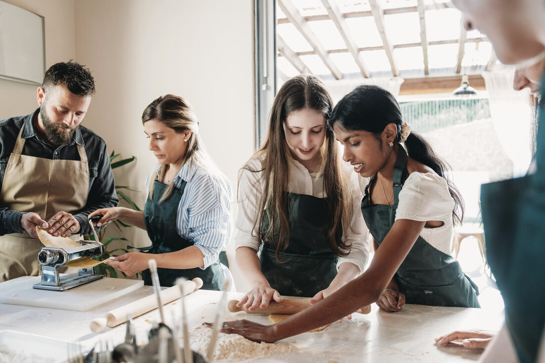 Mehrere Personen arbeiten konzentriert an einer bemehlten Arbeitsfläche: einer dreht Teig durch die Nudelmaschine, zwei andere rollen von Hand aus.