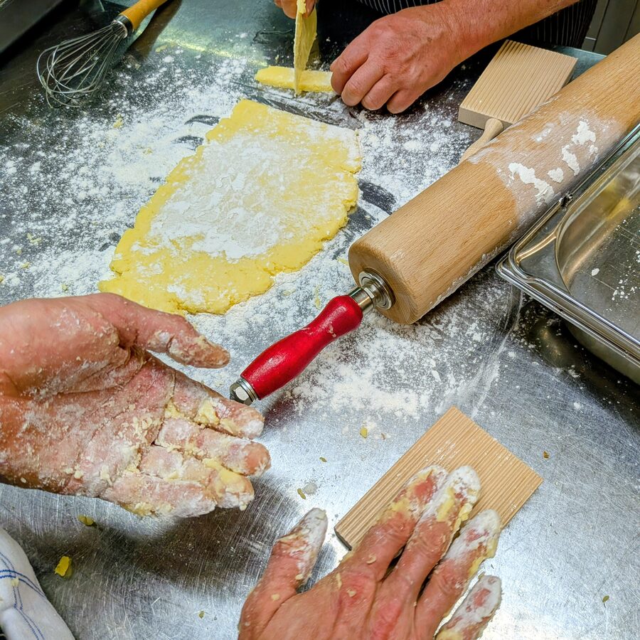 Bemehlte Hände formen auf einem Stahltisch Gnocchi, daneben Wallholz, Gnocchibrett und Schwingbesen.