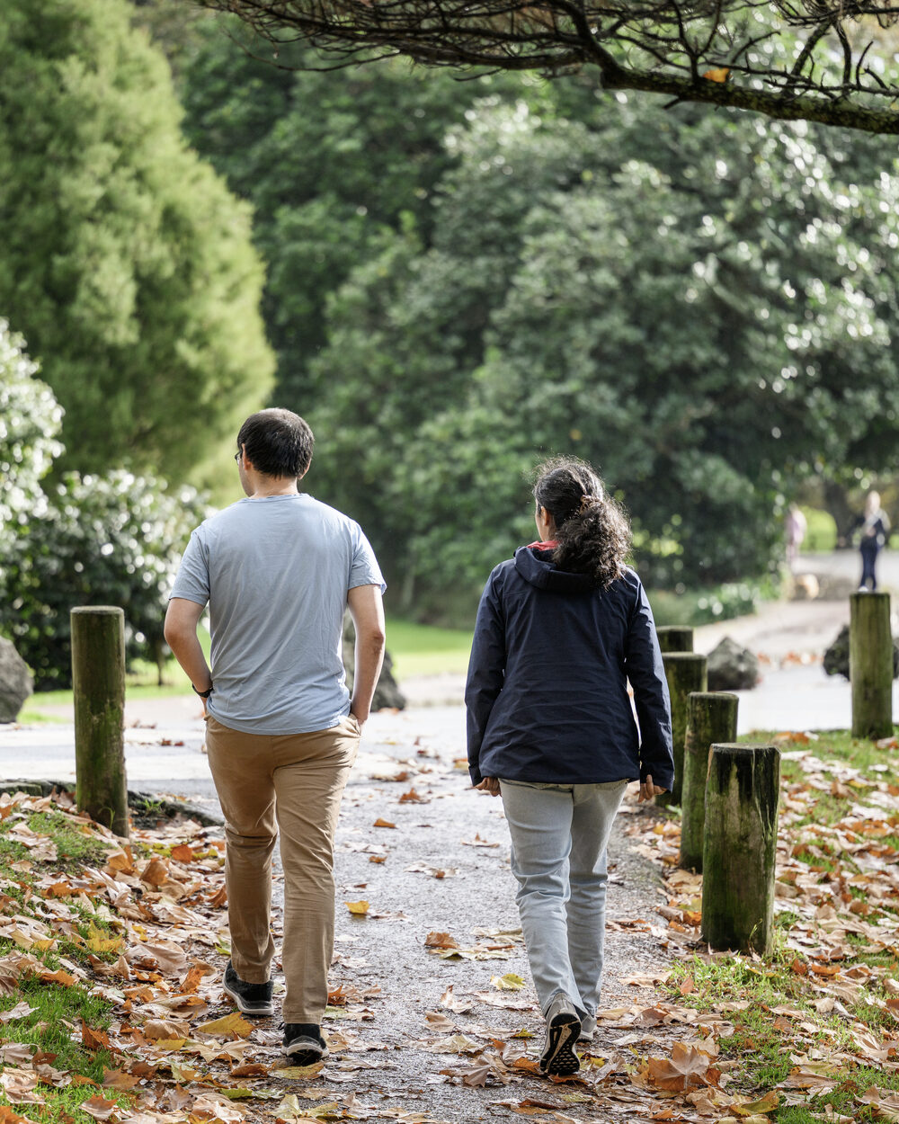 Zwei Personen gehen nebeneinander einen von Laub gesäumten Parkweg entlang, von hinten aufgenommen.