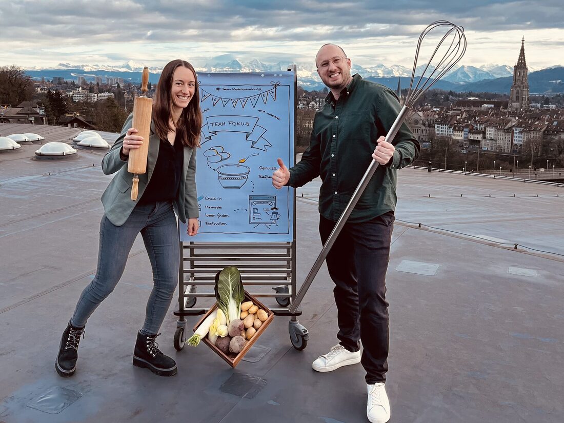 Regula Lenz und Dario Zbinden stehen mit Wallholz und Schwingbesen neben einem Flipchart und einer Harasse Gemüse auf einem Dach über der Stadt Bern.