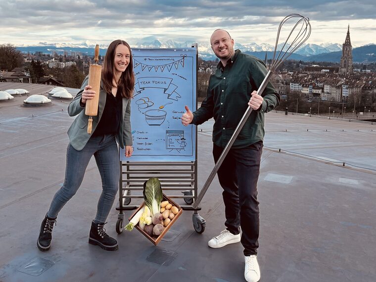 Regula Lenz und Dario Zbinden stehen mit Wallholz und Schwingbesen neben einem Flipchart und einer Harasse Gemüse auf einem Dach über der Stadt Bern.